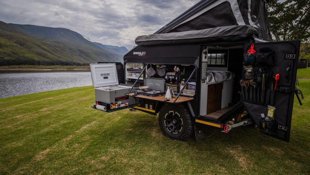 An Infanta 4x4 Enkulu 1 off-road caravan set up with roof up on green grass next to a lake